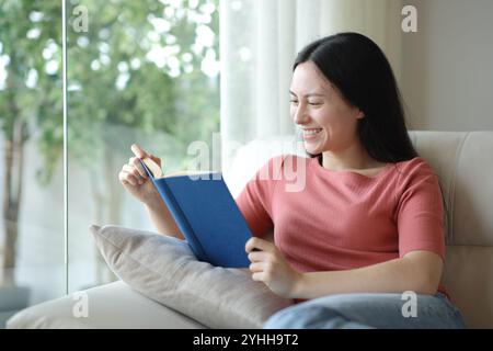 Glückliche asiatische Frau, die ein Papierbuch liest und zu Hause auf einer Couch sitzt Stockfoto