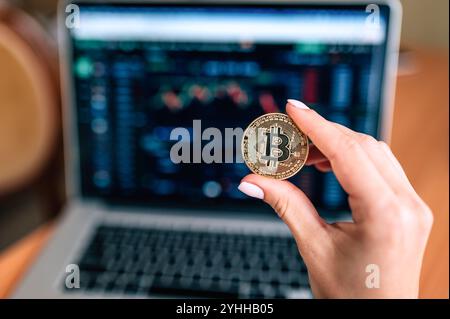 Eine weibliche Hand, die einen goldenen Bitcoin-Münzstempel vor einem Laptop mit Karten hält (von Ivan Radic) Stockfoto