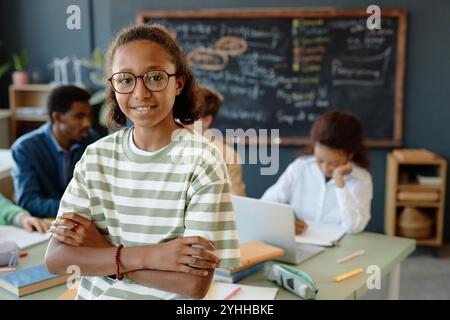 Porträt des Teenagers Afroamerikaner, der eine Brille trägt, während er mit überkreuzten Armen im Schulzimmer steht und in die Kamera lächelt, im Kopierraum Stockfoto