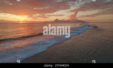 Blick aus der Vogelperspektive auf den hellen orangefarbenen Sonnenuntergang über der Insel Moorea in Französisch-Polynesien. Wellen Rollen sanft das Ufer. Schaffen Sie eine malerische und ruhige Szene mit warmen Farben, die sich auf der Meeresoberfläche spiegeln Stockfoto