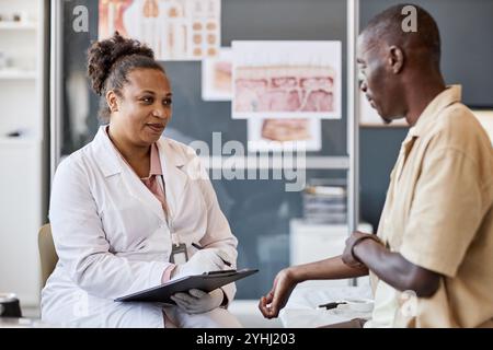 Seitenporträt einer lächelnden Ärztin mit Klemmbrett während der Konsultation des Patienten, der sich über Hautausschlag und Allergien beschwert Stockfoto