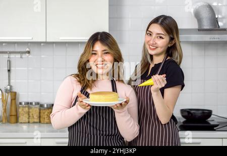 Zwei junge Frauen in Schürze machen Nachtisch in der Küche. Eine Person, die einen Rohrsack hält. Der andere hält Kuchen auf einer weißen Schale. Stockfoto