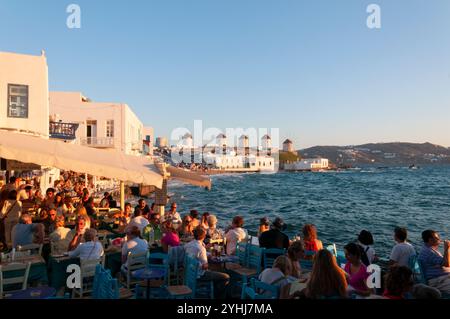 Bar im Freien am Meer mit den berühmten Windmühlen im Hintergrund voll mit Touristen, die bei Sonnenuntergang auf der Insel Mykonos Urlaub machen Stockfoto