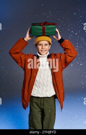 Ein Junge in einem gemütlichen Pullover und einer orangefarbenen Jacke hält glücklich ein Geschenk über dem Kopf im Schnee. Stockfoto