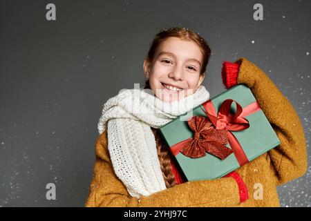 Ein fröhliches Mädchen in warmer Kleidung lächelt und hält ein farbenfrohes Geschenk für Winterfeste. Stockfoto