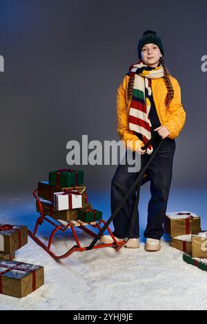 Ein warm gekleidetes Mädchen steht an einem Schlitten voller farbenfroher Weihnachtsgeschenke, bereit für Spaß. Stockfoto