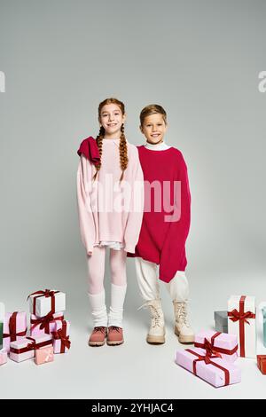 Zwei Kinder in gemütlichen Winteroutfits stehen an farbenfrohen Geschenkboxen voller Jubel. Stockfoto