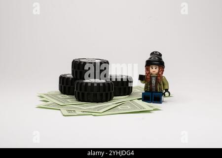 Lego-Mädchen im Winteroutfit, neben Autoreifen gestapelt auf Dollarscheine, isoliert auf weißem, leerem Hintergrund. Stockfoto