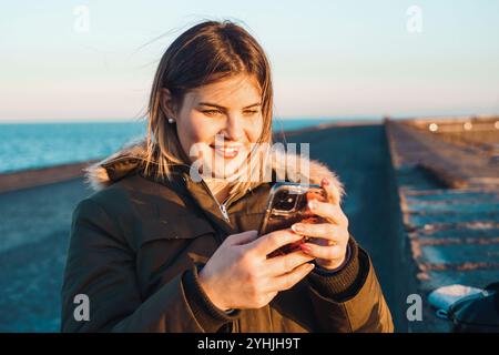 Echte glückliche junge Frau, die während des Sonnenuntergangs am Pier am Meer ein Smartphone benutzt und eine Winterjacke trägt, die die Verbindung zu sozialen Medien genießt Stockfoto