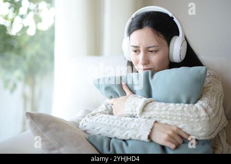 Traurige asiatische Frau, die Kopfhörer trägt und sich beschwert, Audiomaterial zu hören, das Kissen zu Hause umarmt Stockfoto