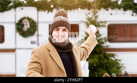 Ein junger Mann genießt während der Winterfeste den Schneefall vor einem gemütlichen Wohnwagen Stockfoto