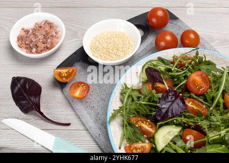 Nahaufnahme von Salat mit frischen Tomaten, Gurken, Rucola, Basilikum, Spinat und Sesam auf dem Teller und dem Schneidebrett mit einem Messer. Schüsseln mit Stockfoto