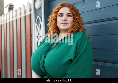 Portrait Plus Size Frau mit lockigem rotem Haar in grüner Kleidung vor bunten blauen hölzernen Wand Hintergrund, Schönheit und Vertrauen im Freien Stockfoto