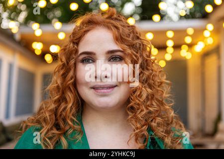 Schöne große Frau mit lockigem rotem Haar lächelt an der Kamera mit unscharfem Bokeh Hintergrund von warmen Lichtern im Freien Abend Stockfoto