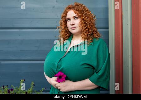 Plus Size Frau mit lockigem rotem Haar in grüner Kleidung hält Blume vor lebendigem blauem Hintergrund, Schönheit und Selbstvertrauen im Freien Stockfoto