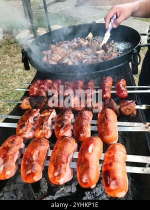 Eine Nahaufnahme mit einer Vielzahl von Fleisch- und Würstchen, die auf einem Grill im Freien brutzeln. Eine Hand wird gesehen, wie sie das Fleisch dreht und das Wesen eines einfängt Stockfoto