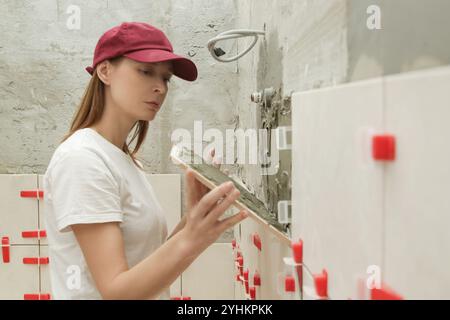 Frau fliesst Badezimmerwände, trägt Klebstoff auf eine Fliese auf und platziert Keramikfliesen in der richtigen Position. Prozess der Installation von Fliesen im Badezimmer Stockfoto