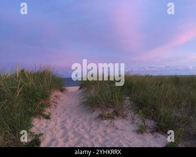 Ein sandiger Pfad, der durch grasbewachsene Dünen zum Strand in der Abenddämmerung führt, mit einem in sanften violetten und rosafarbenen Tönen gemalten Himmel. Die beschauliche Szene fängt den Frieden ein. Stockfoto