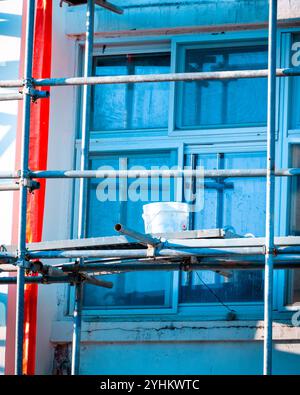 Baugerüst mit rotem Sicherheitsrohr und weißem Eimer vor dem Gebäudefenster Stockfoto