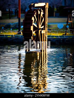 Beleuchtetes hölzernes Wasserrad reflektiert am ruhigen Teich in der Abenddämmerung Stockfoto