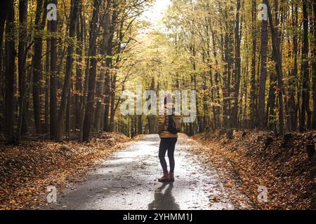 Frau läuft auf einer leeren Landstraße durch einen Herbstwald an sonnigen Tagen. Touristen mit Rucksack Wandern im Wald Stockfoto