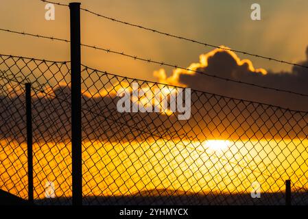 Die Sonne untergeht hinter einem Stacheldrahtzaun und wirft leuchtende Orange- und Gelbtöne über den Himmel, während Wolken über sich ziehen und einen eindrucksvollen Abend A schaffen Stockfoto