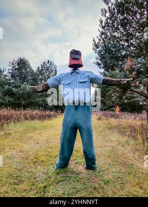 Hausgemachte Vogelscheuche mit einem Plastiktopf für einen Kopf, trägt ein kariertes Hemd, eine aufgesetzte blaue Hose und ausgestreckte Arme auf einer unbefestigten Straße. Stockfoto