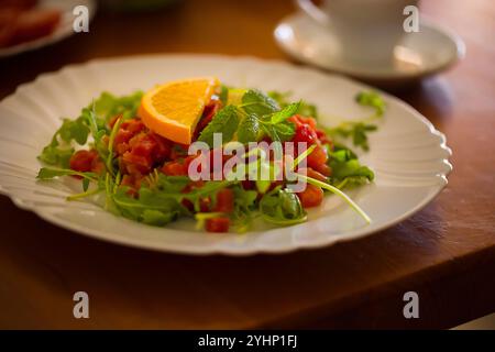 Elegantes Thunfisch-Tatar, garniert mit Rucola und Zitrusfrüchten, sitzt auf einem weißen Teller. Holztisch und Frühstücksgerichte verwischen im Hintergrund sanft Stockfoto