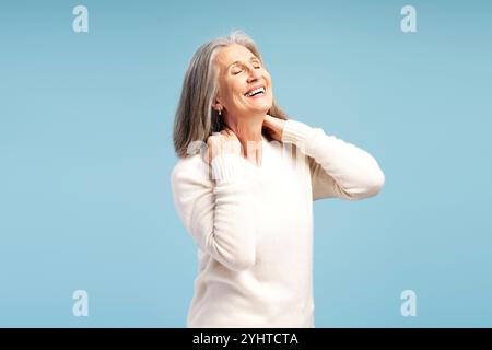 Porträt einer lächelnden älteren grauhaarigen Frau, die den Hals berührt, Schmerzen mit geschlossenen Augen hat, Modemodell, das isoliert auf blauem Hintergrund posiert. Gesundheitswesen Stockfoto