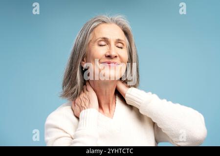 Porträt einer lächelnden älteren Frau, die den Hals berührt, Schmerzen hat, reife Frau, die sich entspannt, isoliert auf blauem Hintergrund steht. Behandlungskonzept Stockfoto