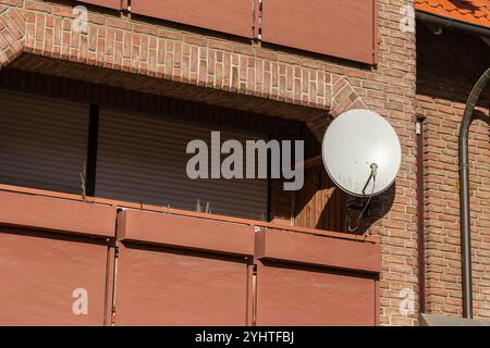 An einem sonnigen Tag befindet sich eine große Satellitenschüssel an der Außenseite eines Backsteingebäudes, neben einem geschlossenen Fenster mit Holzläden. Stockfoto