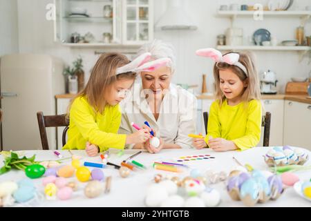 Frohe Ostern. Zwei kleine Schwestern Zwillinge, Großmutter, die Eier malt und die gemeinsame Zeit genießt. Glückliche Familie Großmutter Enkelinnen Kinder bereiten sich auf Ostern vor. Christliche Festivaltradition des Frühlings Stockfoto