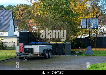 Ein Basketballkorb steht in einem Hinterhof neben einem geparkten Anhänger und bunten Mülltonnen, umgeben von Grün und Bäumen an einem sonnigen Herbsttag. Stockfoto