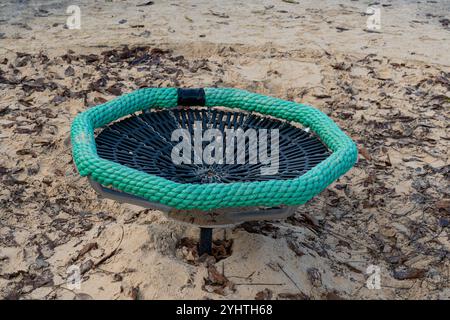 In einem Sandbereich befindet sich ein runder Spielplatz im Freien mit einem gewebten Korbdesign mit grünem Seilrand. Es verschmilzt in ein natürliches, lea Stockfoto