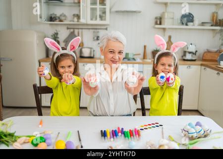 Frohe Ostern. Zwei kleine Schwestern Zwillinge, Großmutter, die Eier malt und die gemeinsame Zeit genießt. Glückliche Familie Großmutter Enkelinnen Kinder bereiten sich auf Ostern vor. Christliche Festivaltradition des Frühlings Stockfoto