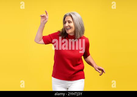 Porträt einer attraktiven älteren grauhaarigen Frau, die einen roten Pullover trägt und Spaß auf gelbem Hintergrund hat. Hobby-Konzept Stockfoto