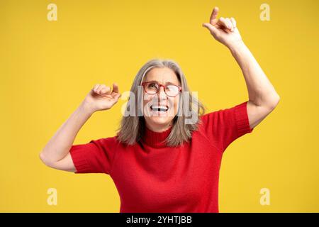 Porträt lächelnde ältere Frau mit stilvoller Brille und rotem Pullover, der isoliert auf gelbem Hintergrund tanzt. Unterhaltsames Konzept Stockfoto