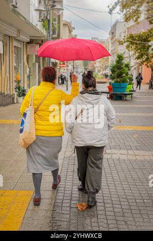 Zwei Frauen laufen unter einem hellroten Schirm in einer regnerischen Stadtstraße Stockfoto