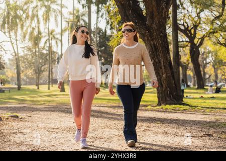 Zwei Frauen spazieren in einem Stadtpark, lächeln und plaudern. Die warme Nachmittagssonne unterstreicht das Grün und die entspannte Atmosphäre beim Spaziergang Stockfoto