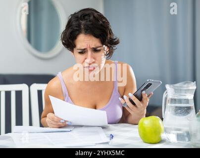Schockierte junge Frau, die Dokumente ansieht und Smartphone in der Hand hält Stockfoto