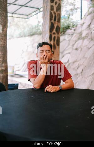 Ein Mann in einem roten T-Shirt sitzt nachdenklich an einem schwarzen Tisch und legt sein Kinn auf seine Hand. Die Umgebung im Freien mit Steinwänden und natürlichem Licht ergänzen Stockfoto