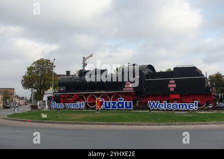 Historische schwarze Dampflokomotive auf einem Kreisverkehr mit Begrüßungsschildern in Buzău, Rumänien, unter bewölktem Himmel Stockfoto