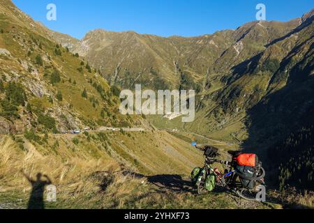 Fahrräder mit Tourenausrüstung ruhen auf einem malerischen Berghang mit Blick auf eine kurvenreiche Straße und das zerklüftete Alpental und den Schatten der Radfahrer Stockfoto