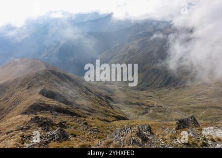 Neblige Berglandschaft mit zerklüfteten Gipfeln und grasbewachsenen Hängen, die teilweise von Wolken bedeckt sind, an einem dramatischen bewölkten Tag aus Moldoveanu, Rumänien Stockfoto