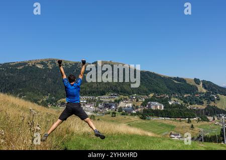 Man in blue shirt jumps joyfully on a hillside with stunning view of a Donovaly ski town in Slovakia under a clear summer blue sky Stockfoto