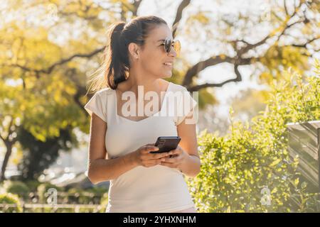 Hübsche junge Frau mit Sonnenbrille, weißem Muskelhemd und rosa jean besucht ihr Smartphone in einem sonnendurchfluteten Park, umgeben von Grün. Stockfoto