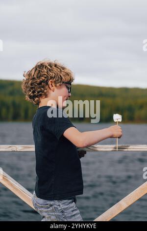 Niedlicher Junge mit blonden lockigen Haaren, der am See einen gerösteten Marshmallow auf einem Spieß genießt, während er am See steht und sich in der Natur mit purer Freude im Gesicht zeigt Stockfoto