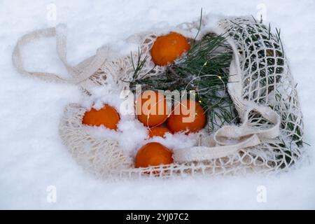 Reife Mandarinen im wiederverwendbaren Netzbeutel mit Tannenzweig im verschneiten Winter. Konzept nachhaltiger Weihnachtsfeier, Verantwortung und Inklusivität. Orangen im Baumwollbeutel Stockfoto