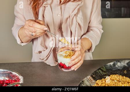 Eine Person fügt einem Parfait in einem Glas eine Krümelschicht hinzu, wodurch eine knusprige Textur entsteht, die die cremigen und fruchtigen Schichten ergänzt. Stockfoto