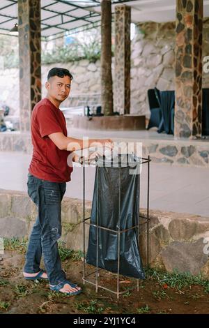 Ein Mann in rotem Hemd und Jeans entsorgt den Abfall im Freien in einem schwarzen Plastikmüll und fördert so eine ordnungsgemäße Abfallentsorgung in einer sauberen Umgebung. Stockfoto